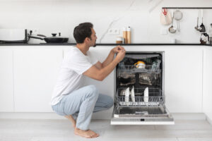 A man kneeling in front of a dishwasher, fixing its door.
