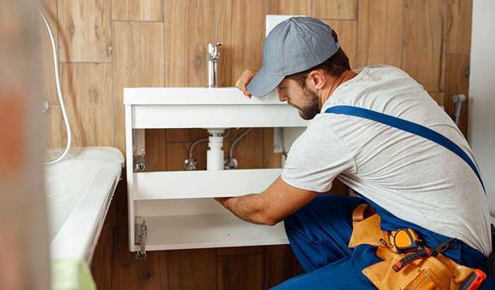 A man in a blue shirt and hat fixing a sink.
