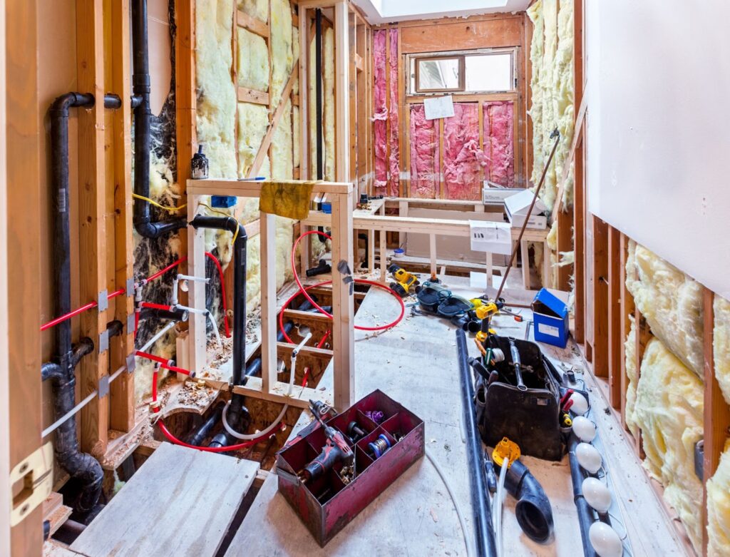 Construction site with exposed wooden framing, insulation, plumbing pipes, and tools scattered around.