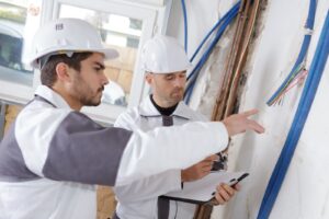 Two workers in hard hats inspecting electrical wiring on a wall.