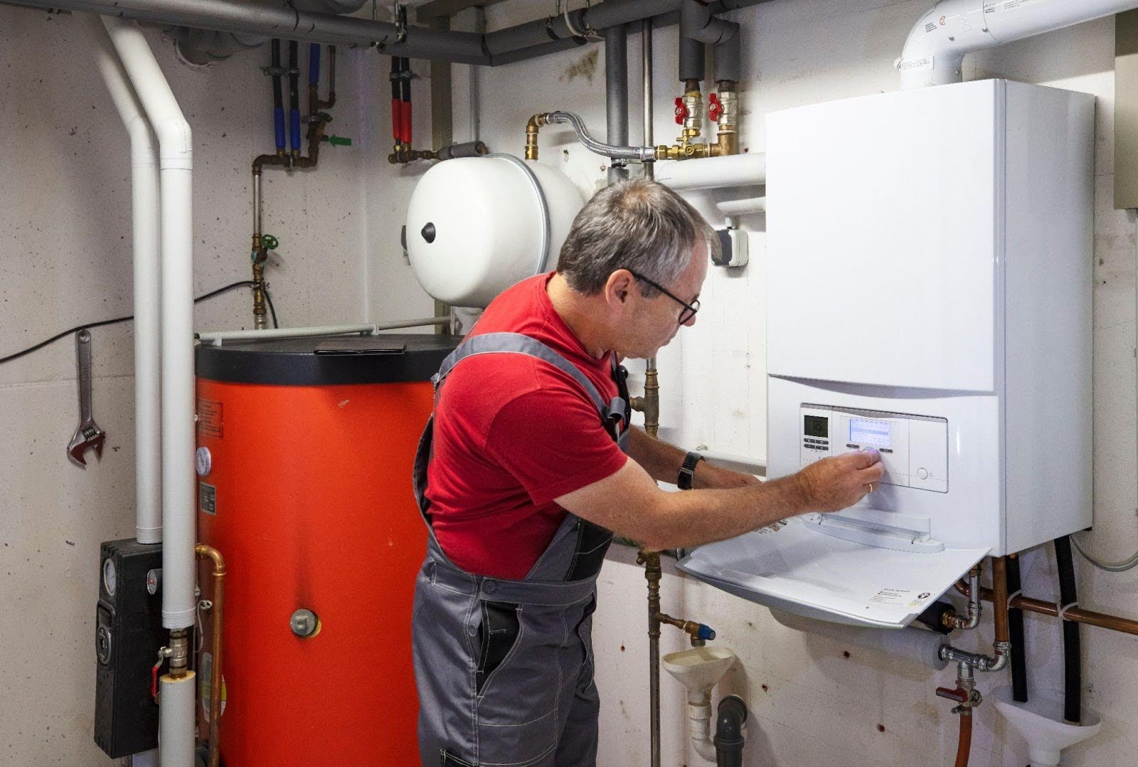 A man in a red shirt and pants is repairing a gas boiler focused on his work in a well-lit utility room