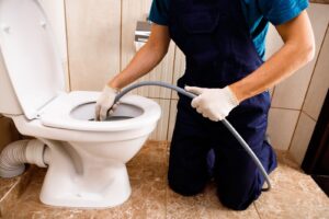 Plumber in blue overalls and gloves using a drain snake to unclog a white toilet in a tiled bathroom.